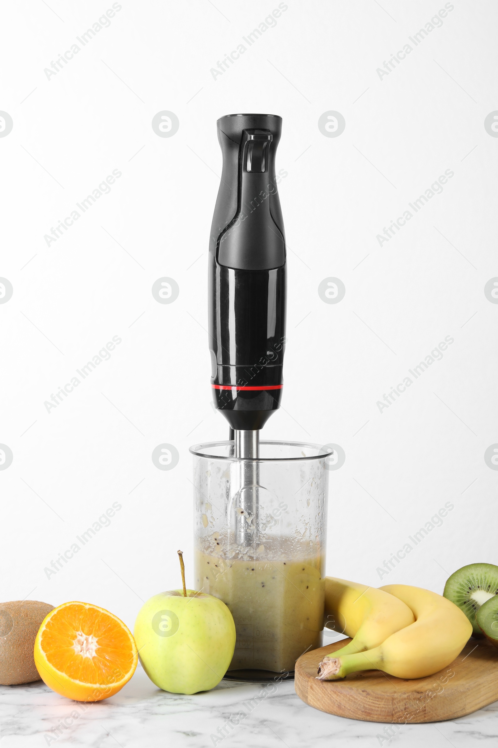 Photo of Hand blender with mixture of ingredients and fresh fruits on white marble table