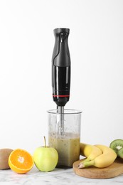 Photo of Hand blender with mixture of ingredients and fresh fruits on white marble table