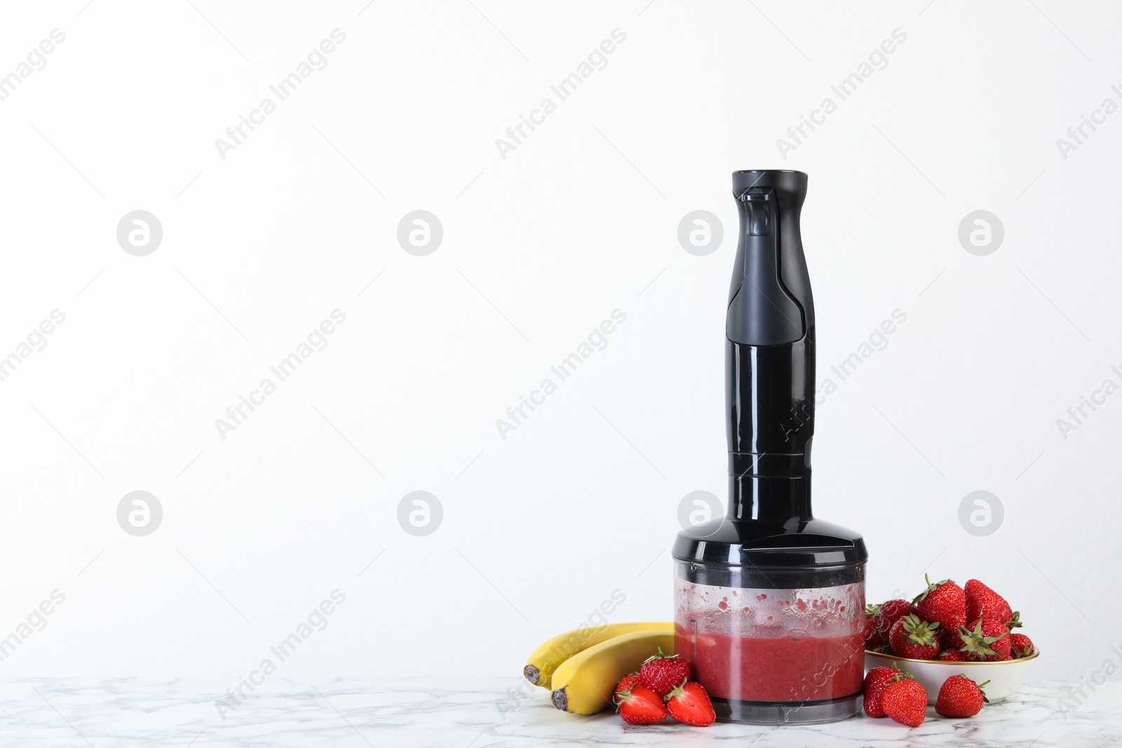 Photo of Hand blender with mixture of ingredients and fresh fruits on white marble table, space for text