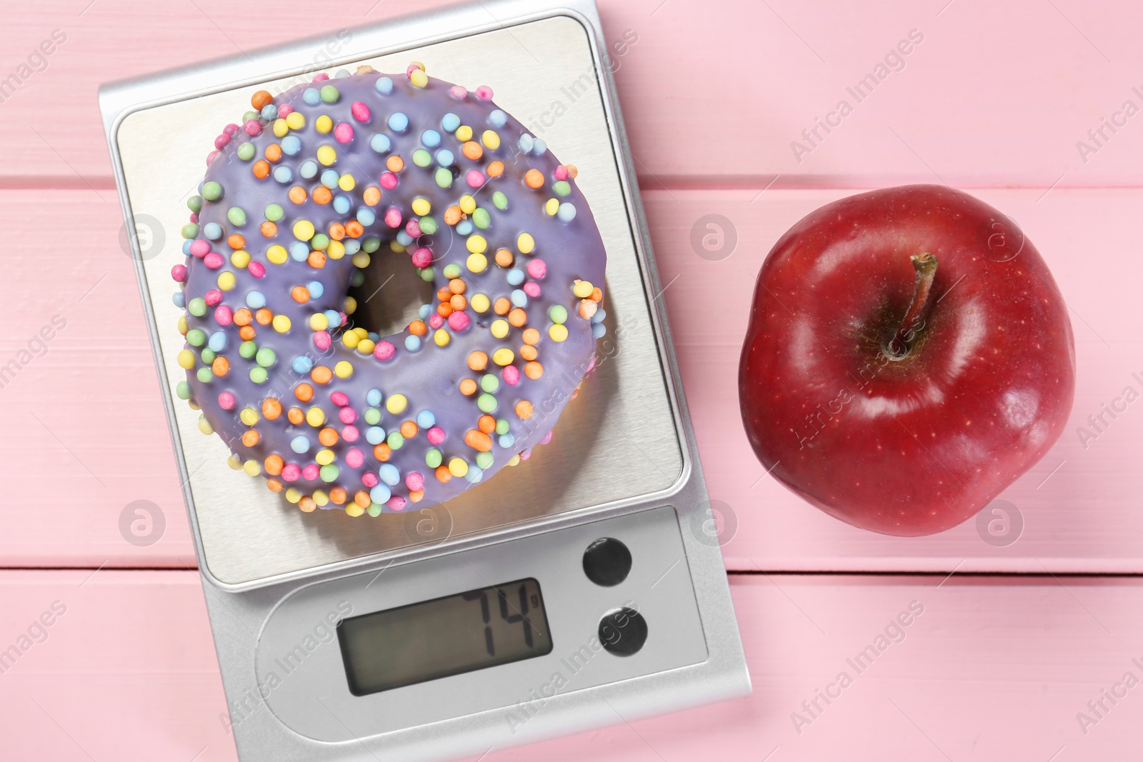 Photo of Kitchen scale with donut and apple on pink wooden table, top view