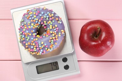 Photo of Kitchen scale with donut and apple on pink wooden table, top view