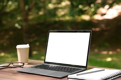 Photo of Laptop, notebook, pen, glasses and paper cup on wooden table outdoors. Remote work