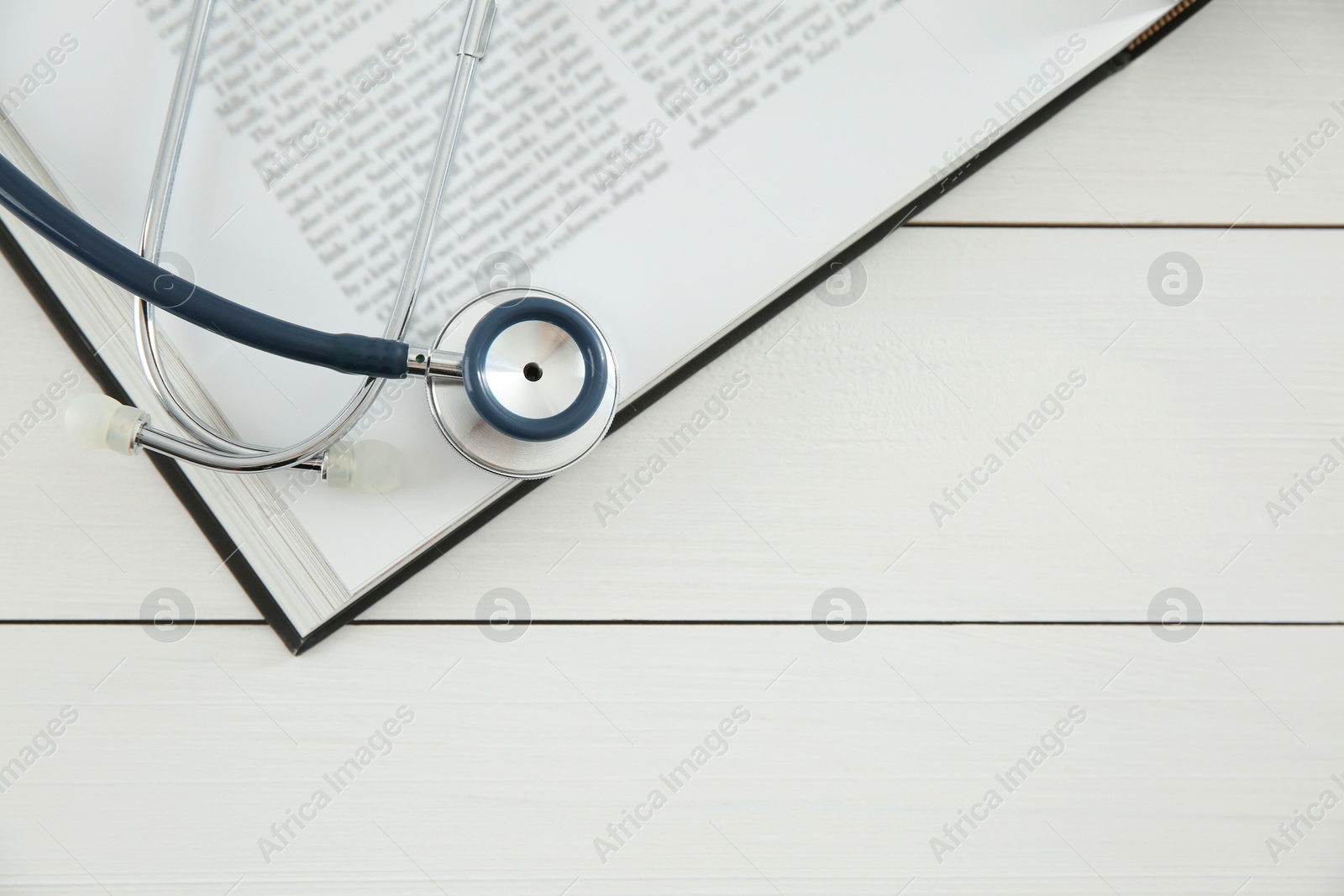 Photo of One new medical stethoscope and book on white wooden table, top view. Space for text