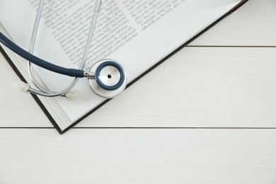 One new medical stethoscope and book on white wooden table, top view. Space for text