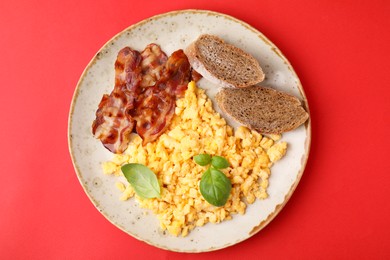 Photo of Delicious scrambled eggs with bacon and basil in plate on red background, top view