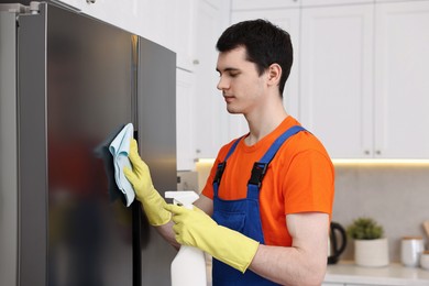 Photo of Professional janitor wearing uniform cleaning fridge in kitchen