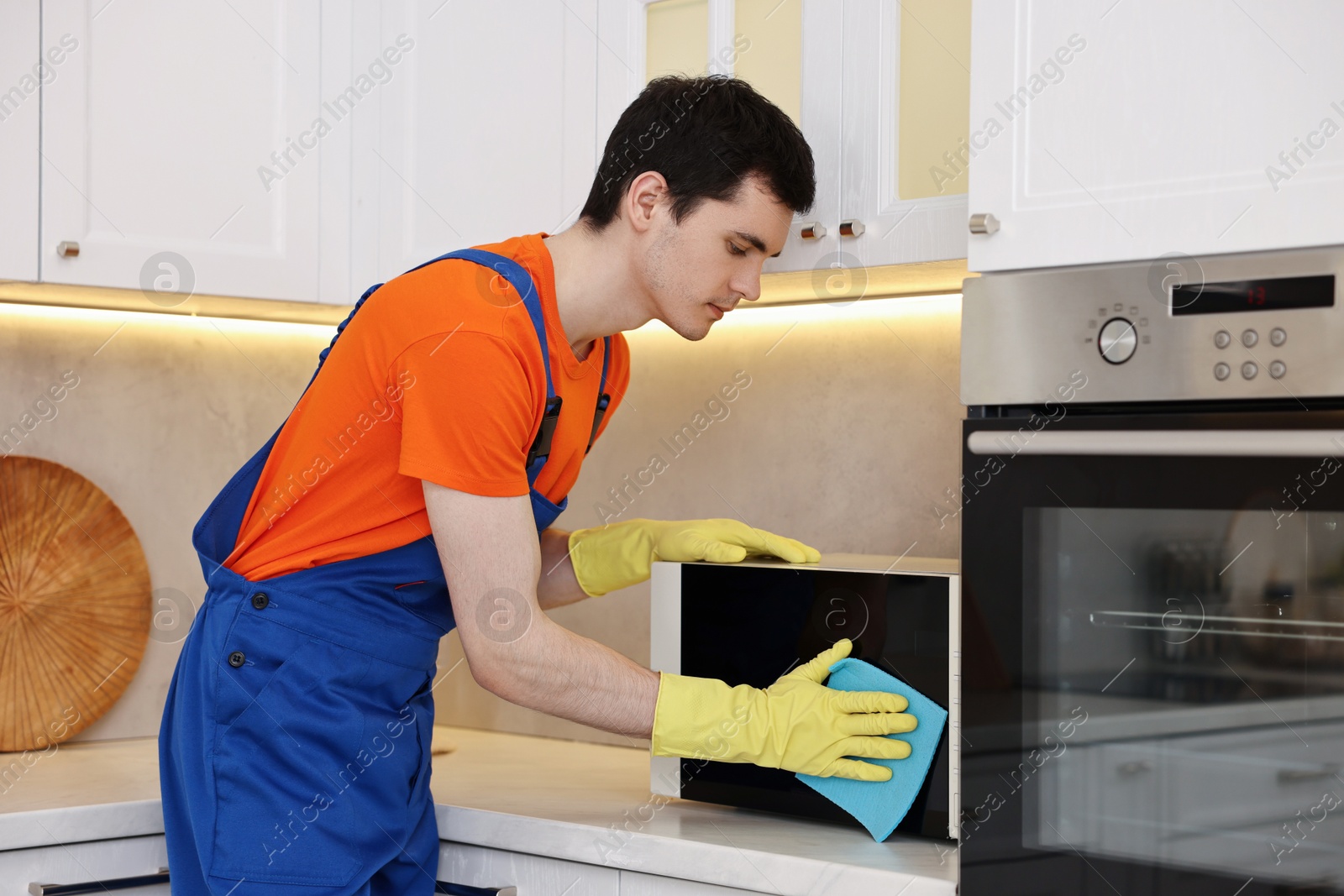 Photo of Professional janitor wearing uniform cleaning microwave oven in kitchen