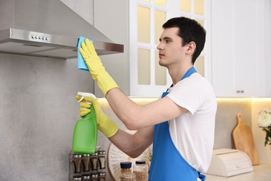 Photo of Professional janitor wearing uniform cleaning kitchen hood with rag and detergent indoors