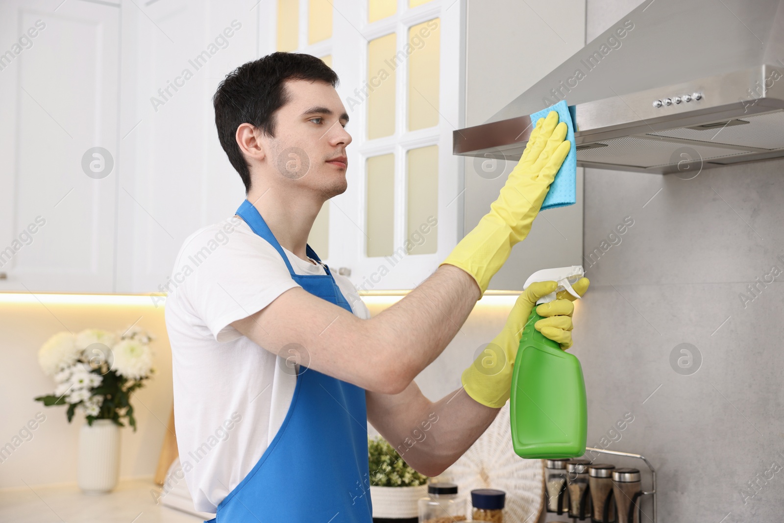 Photo of Professional janitor wearing uniform cleaning kitchen hood with rag and detergent indoors