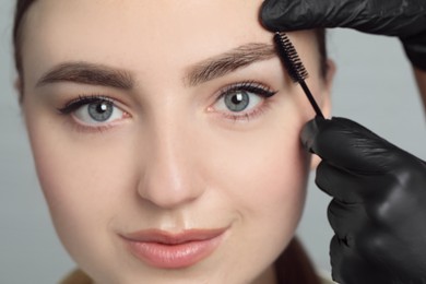 Photo of Beautician brushing young woman's eyebrow on light background, closeup