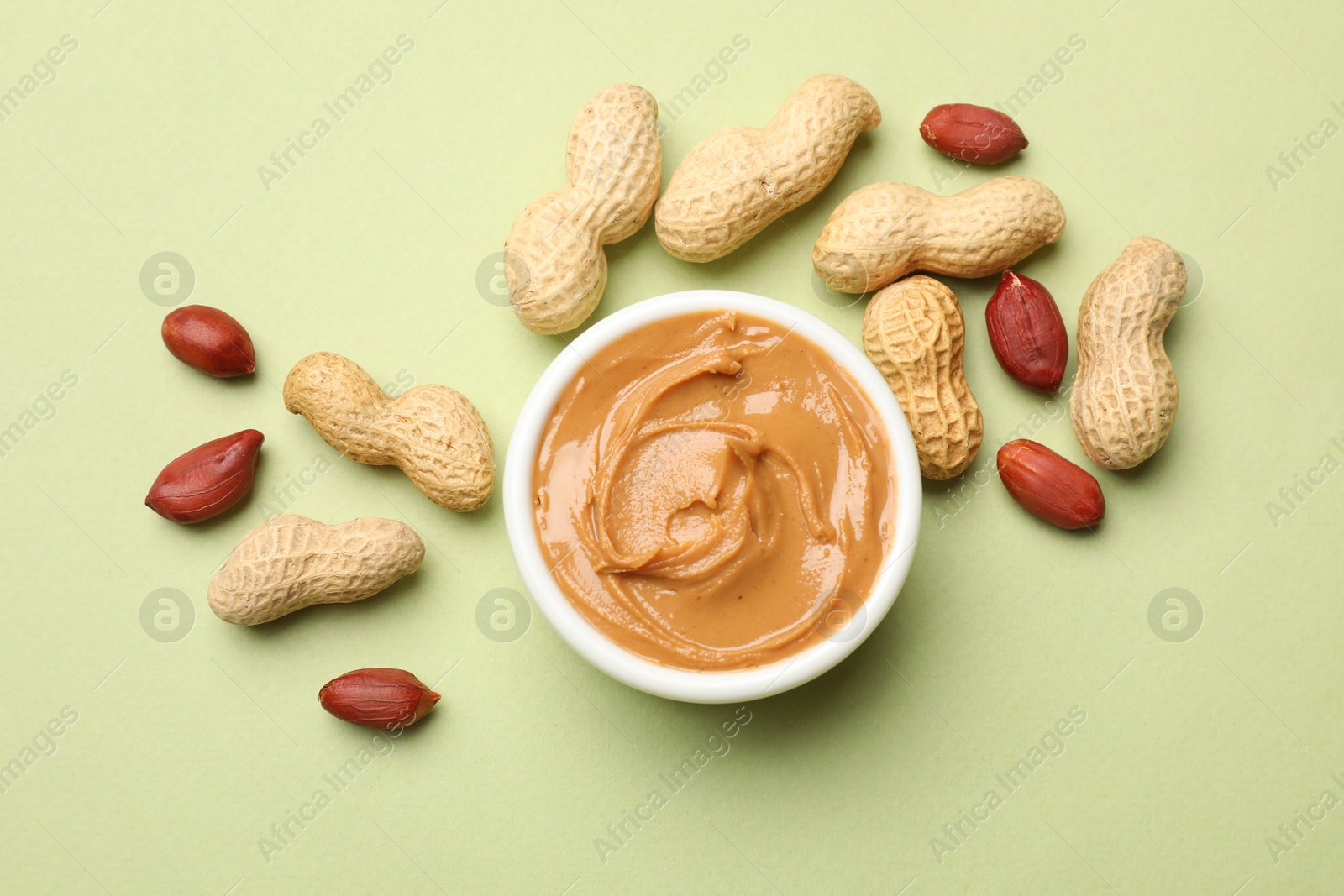 Photo of Tasty peanut butter in bowl and groundnuts on pistachio color background, flat lay