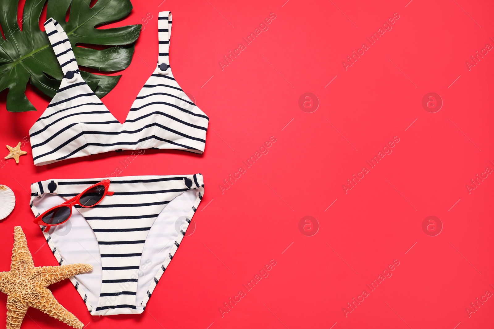 Photo of Striped swimsuit, sunglasses, starfishes and monstera leaf on red background, flat lay. Space for text