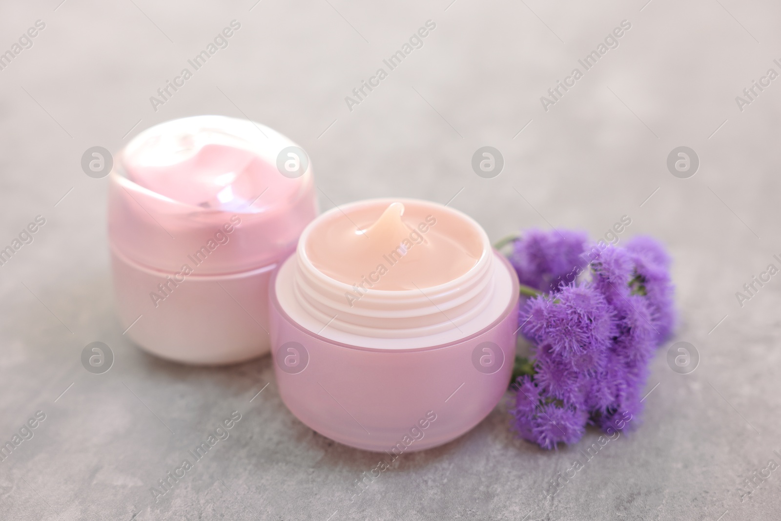 Photo of Jars of creams and ageratum flowers on gray table, closeup