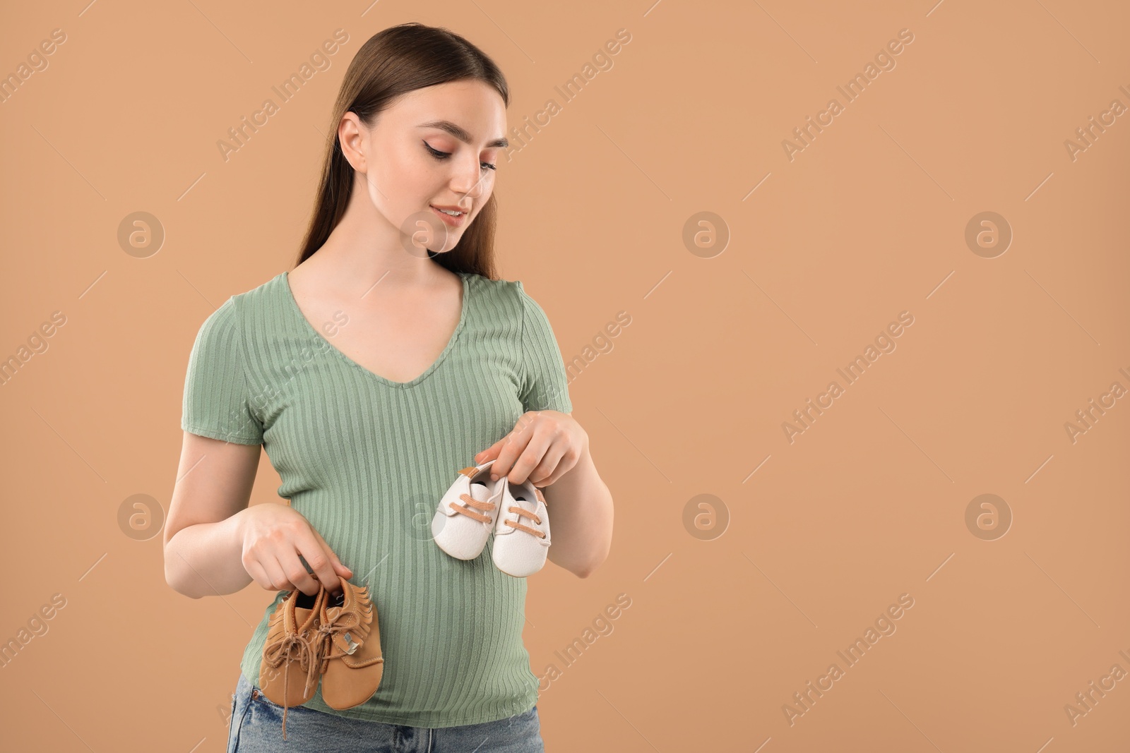 Photo of Expecting twins. Pregnant woman holding two pairs of shoes on light brown background, space for text