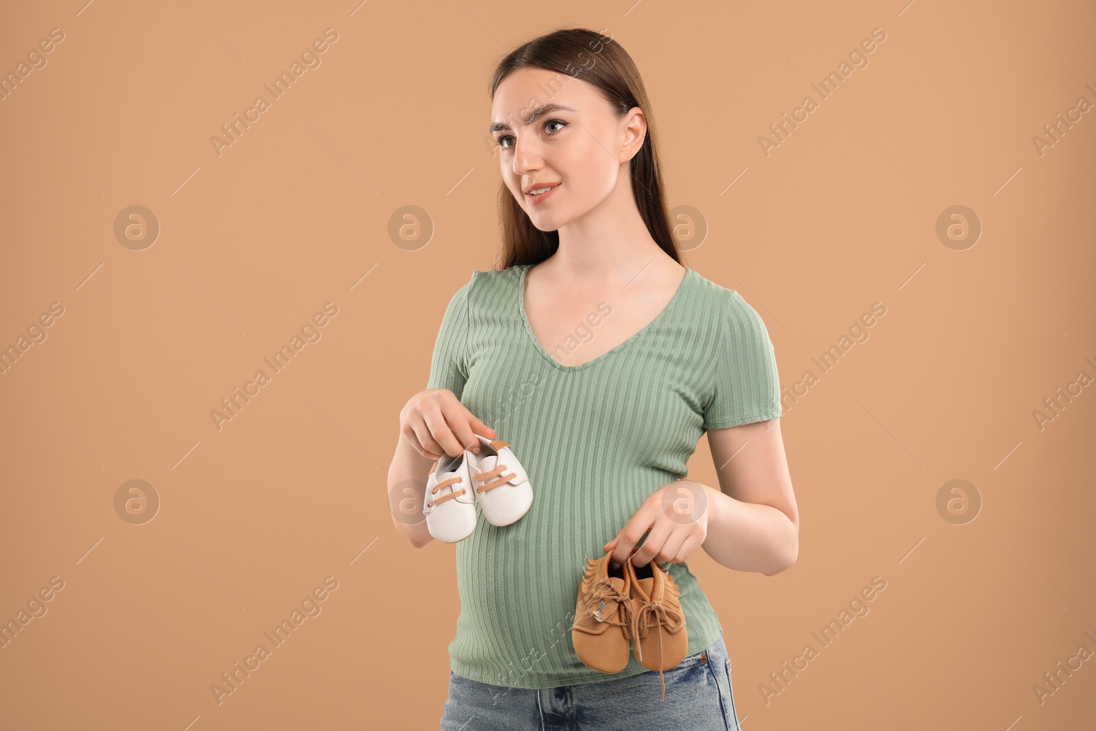 Photo of Expecting twins. Pregnant woman holding two pairs of shoes on light brown background