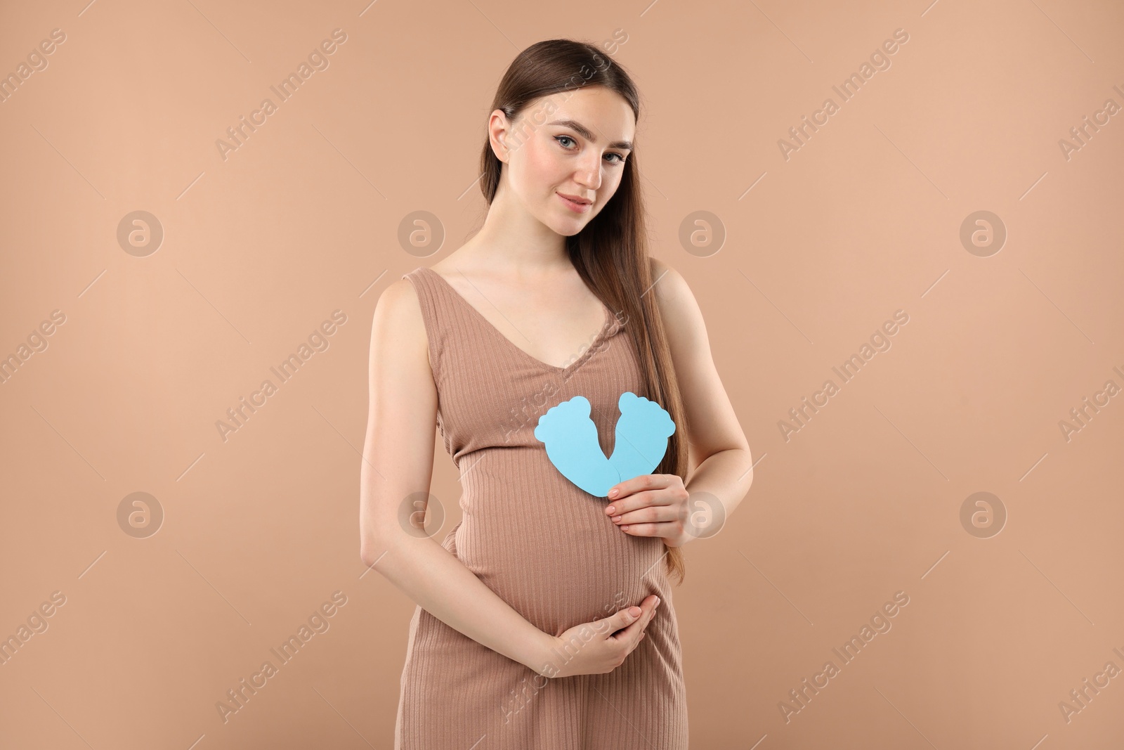 Photo of Expecting twins. Pregnant woman holding two paper cutouts of feet on light brown background