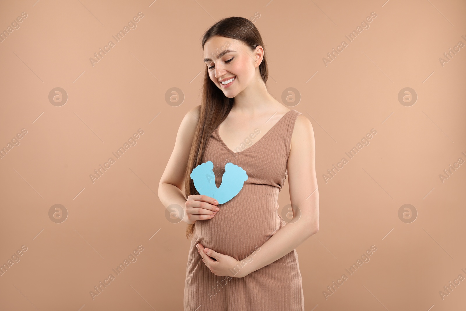 Photo of Expecting twins. Pregnant woman holding two paper cutouts of feet on light brown background