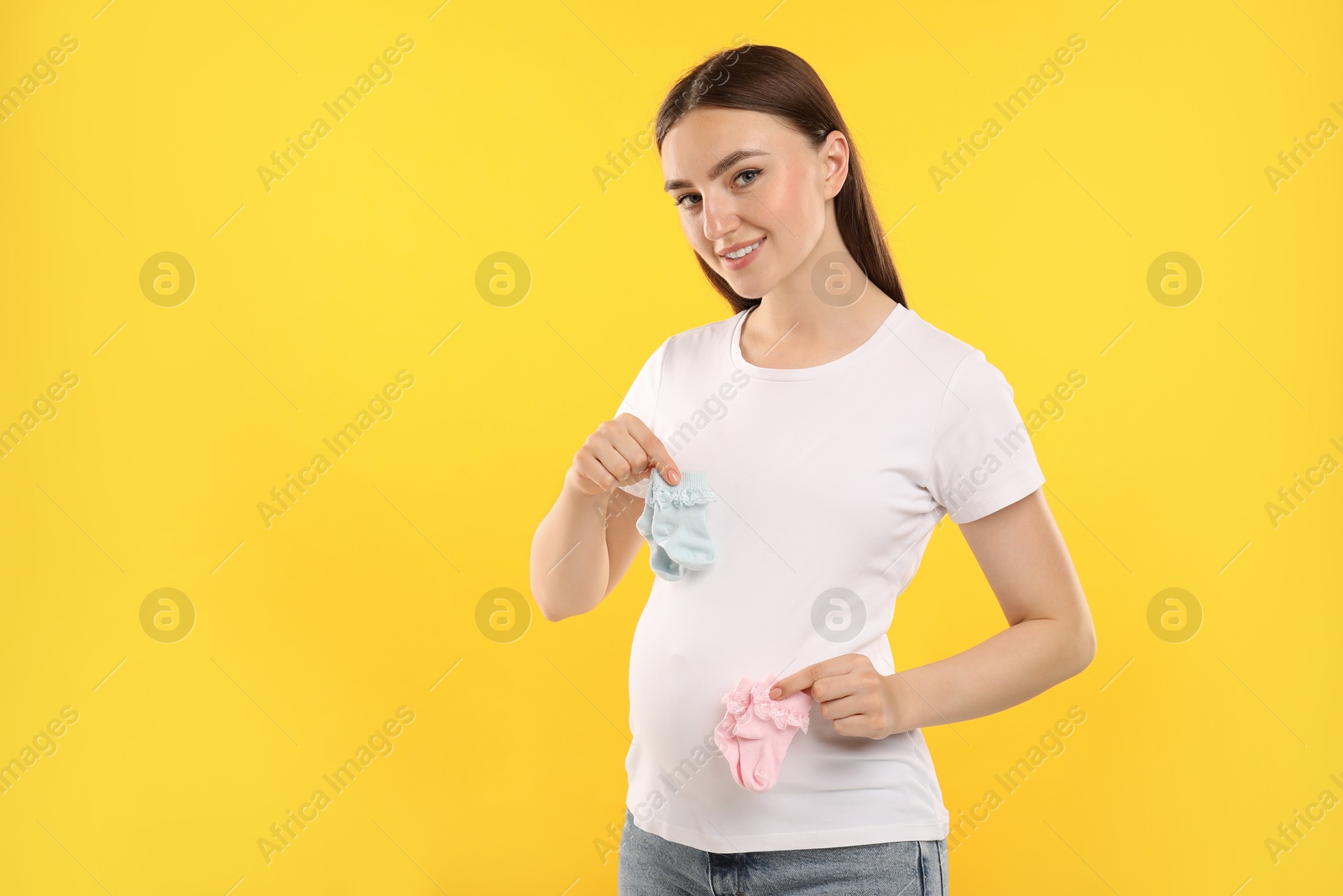 Photo of Expecting twins. Pregnant woman holding two pairs of socks on yellow background, space for text