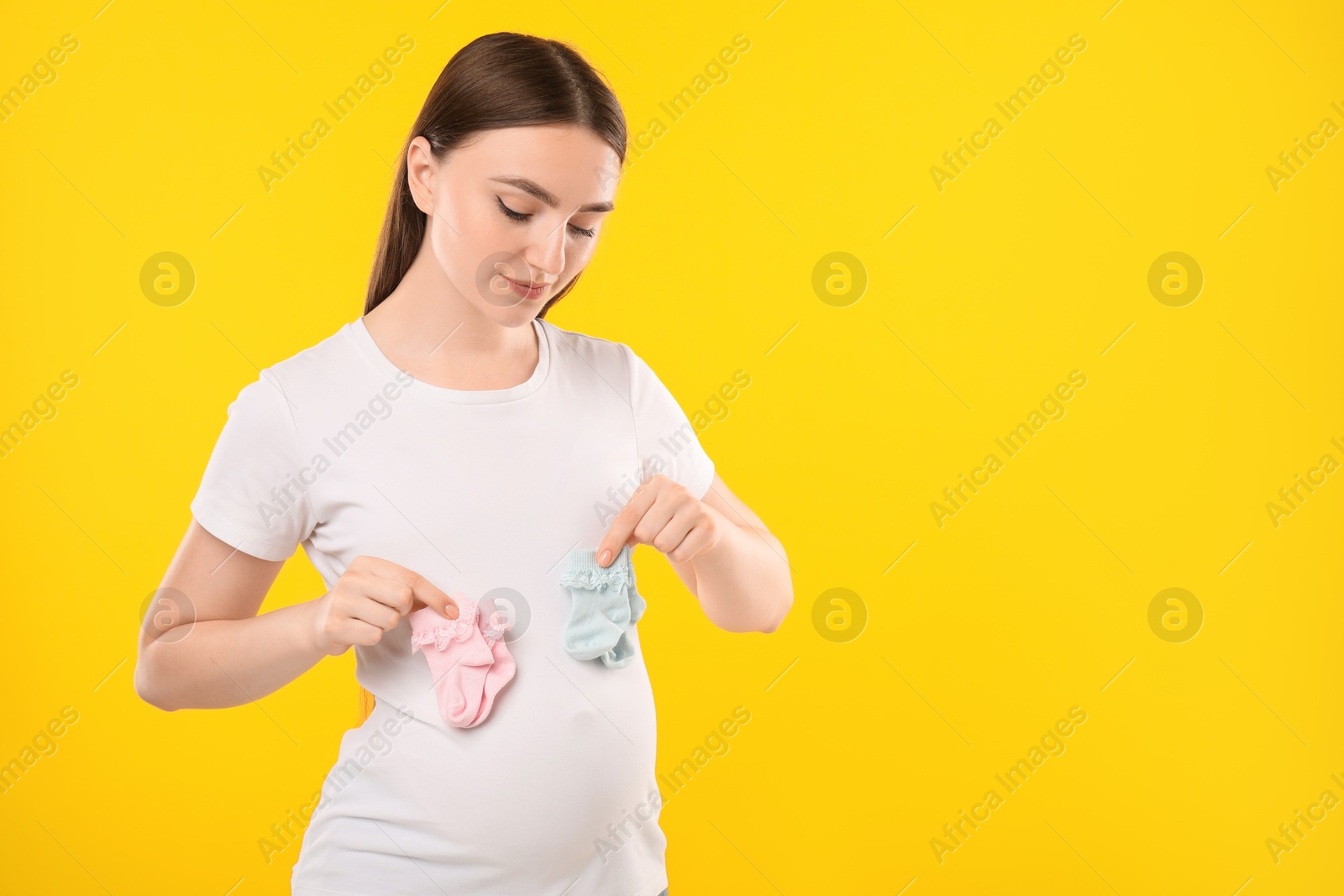 Photo of Expecting twins. Pregnant woman holding two pairs of socks on yellow background, space for text