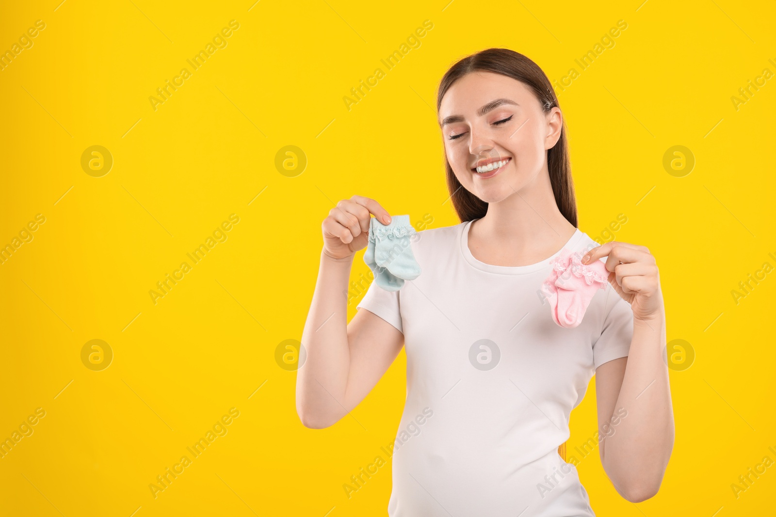 Photo of Expecting twins. Pregnant woman holding two pairs of socks on yellow background, space for text