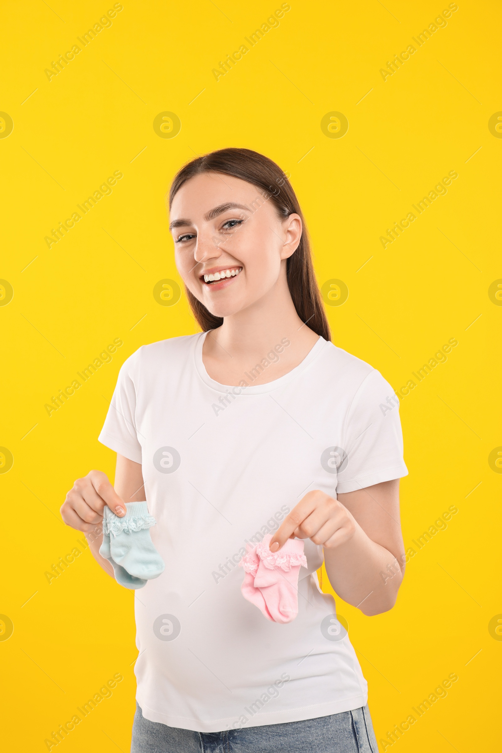 Photo of Expecting twins. Pregnant woman holding two pairs of socks on yellow background