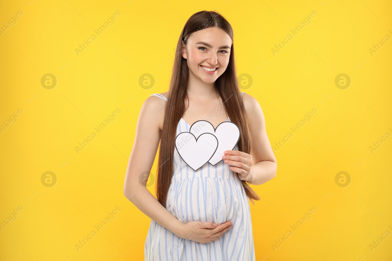 Photo of Expecting twins. Pregnant woman holding two paper cutouts of hearts on yellow background