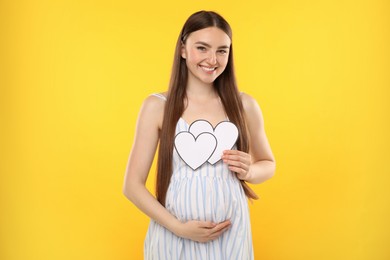 Photo of Expecting twins. Pregnant woman holding two paper cutouts of hearts on yellow background