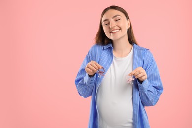 Photo of Expecting twins. Pregnant woman holding two pacifiers on pink background, space for text