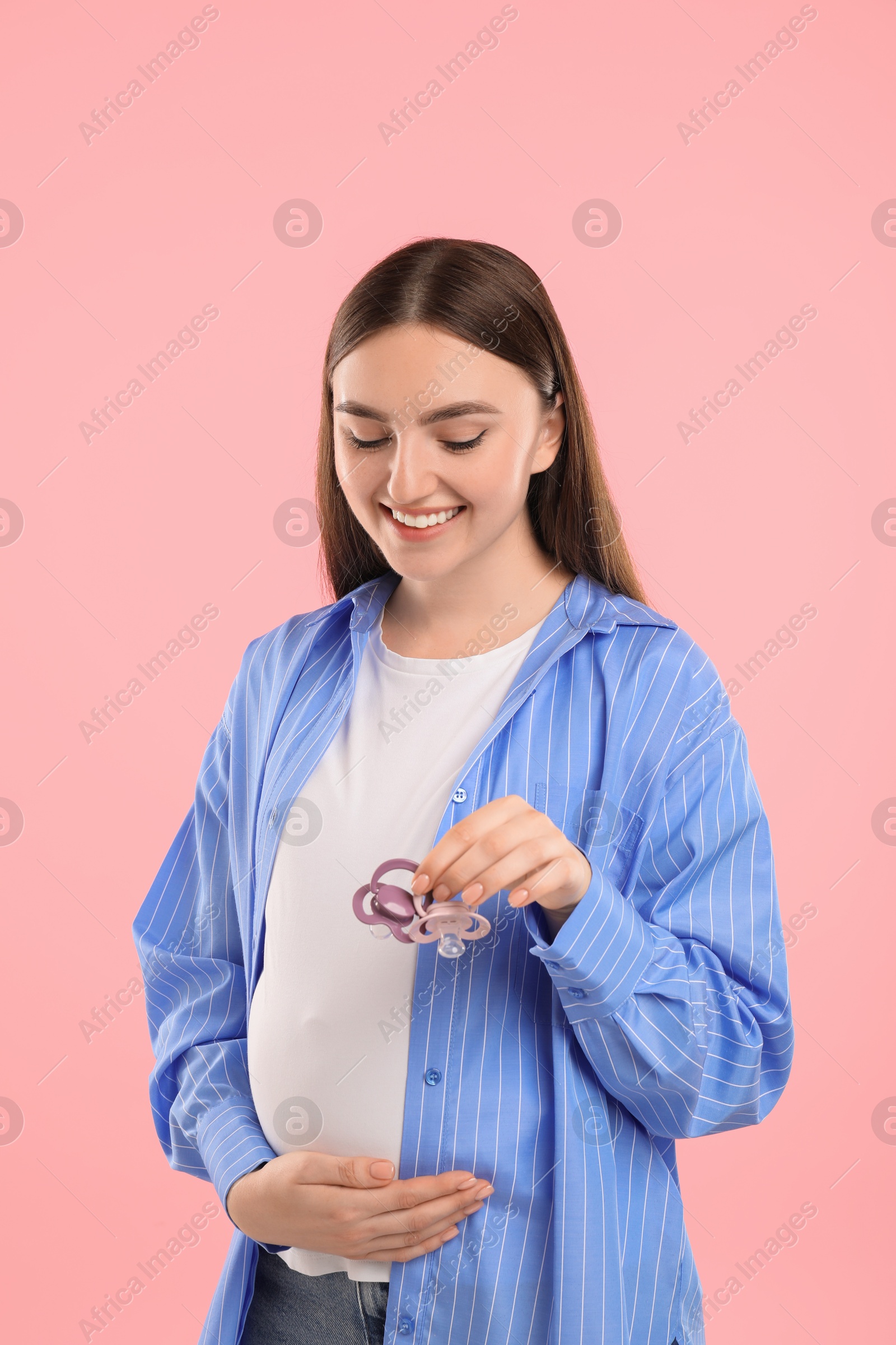 Photo of Expecting twins. Pregnant woman holding two pacifiers on pink background