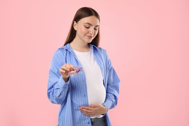 Photo of Expecting twins. Pregnant woman holding two pacifiers on pink background, space for text