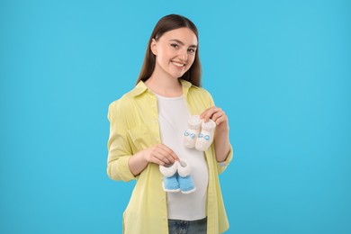 Photo of Expecting twins. Pregnant woman holding two pairs of baby shoes on light blue background