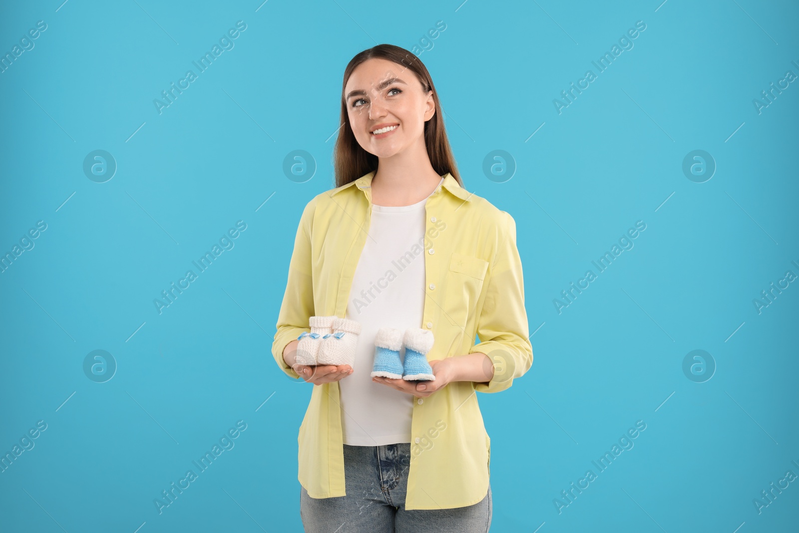 Photo of Expecting twins. Pregnant woman holding two pairs of baby shoes on light blue background