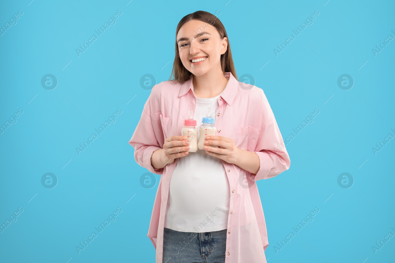 Photo of Expecting twins. Pregnant woman holding two bottles with milk on light blue background