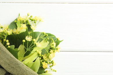 Fresh linden leaves and flowers in bag on white wooden table, top view. Space for text