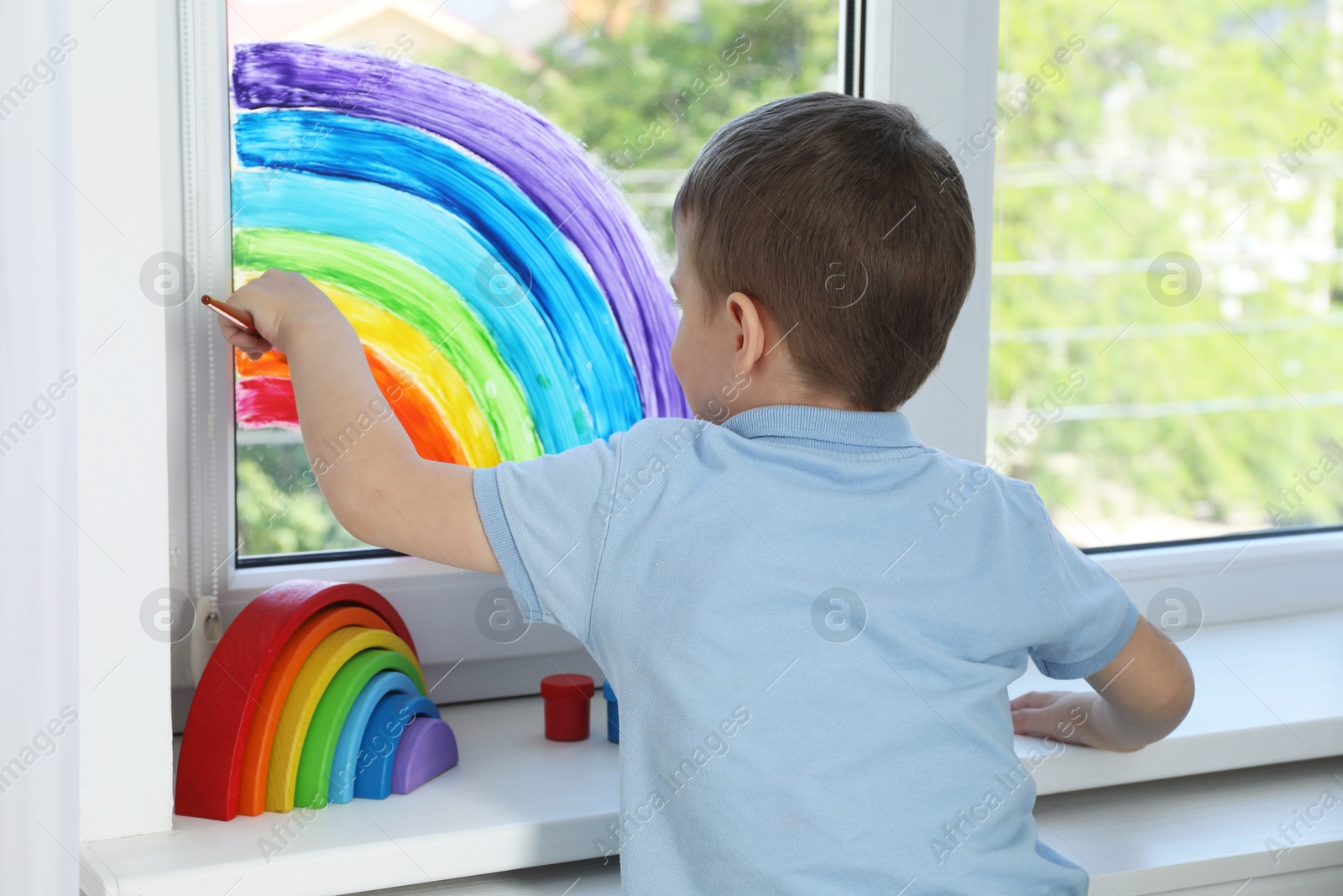 Photo of Little boy drawing rainbow on window indoors