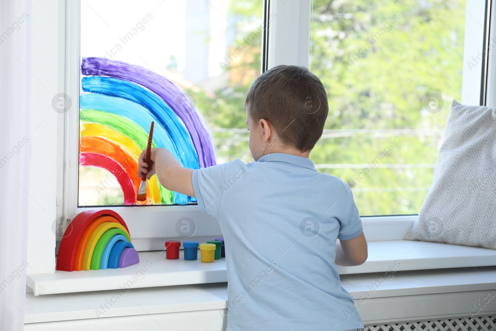 Photo of Little boy drawing rainbow on window indoors