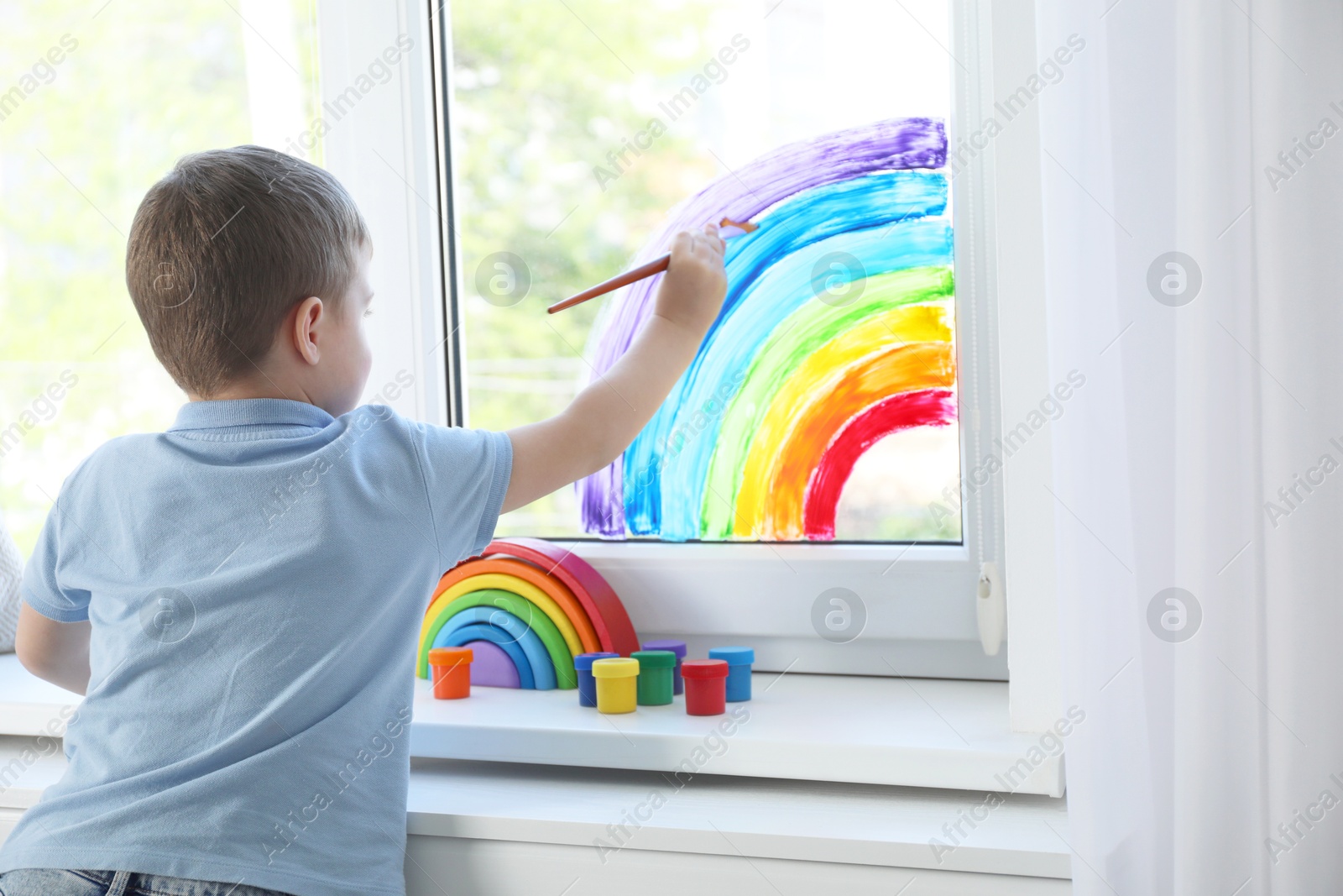 Photo of Little boy drawing rainbow on window indoors