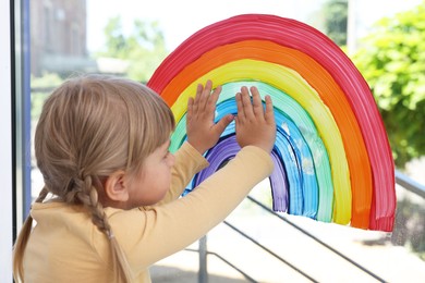 Photo of Little girl touching picture of rainbow on window indoors