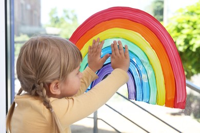 Little girl touching picture of rainbow on window indoors