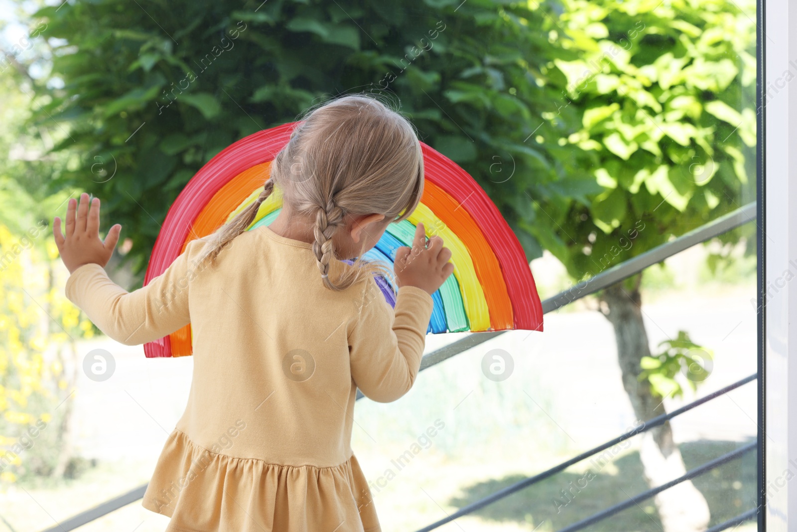 Photo of Little girl touching picture of rainbow on window indoors, back view