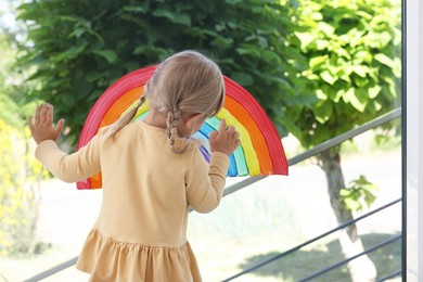 Photo of Little girl touching picture of rainbow on window indoors, back view