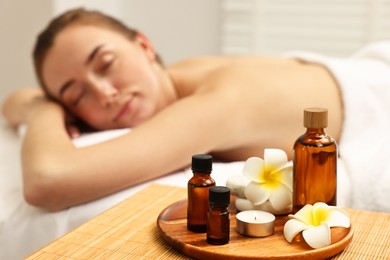 Photo of Aromatherapy. Woman relaxing on massage couch in spa salon, focus on bottles of essential oils, burning candle and plumeria flowers