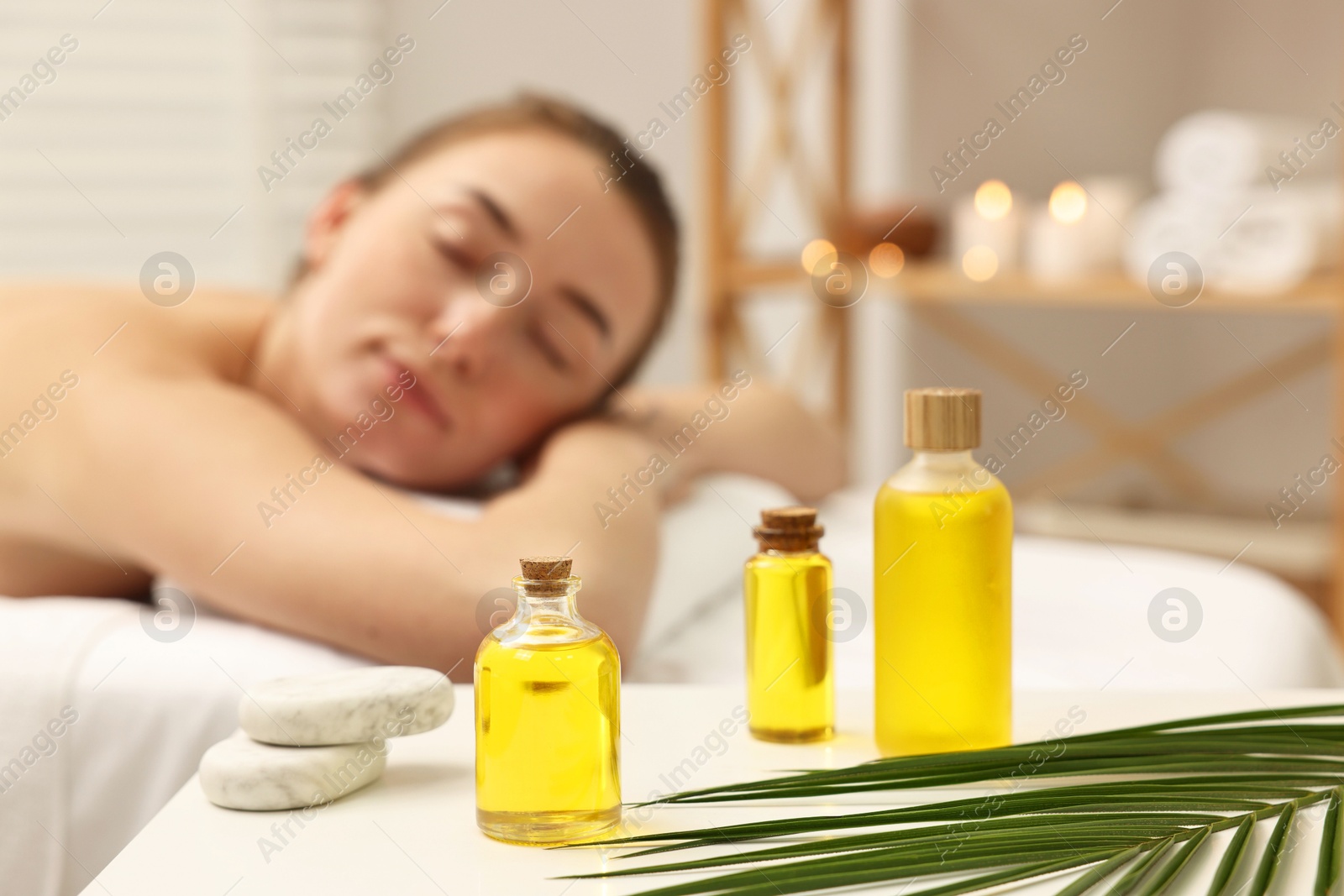 Photo of Aromatherapy. Woman relaxing on massage couch in spa salon, focus on bottles of essential oils, stones and palm leaf