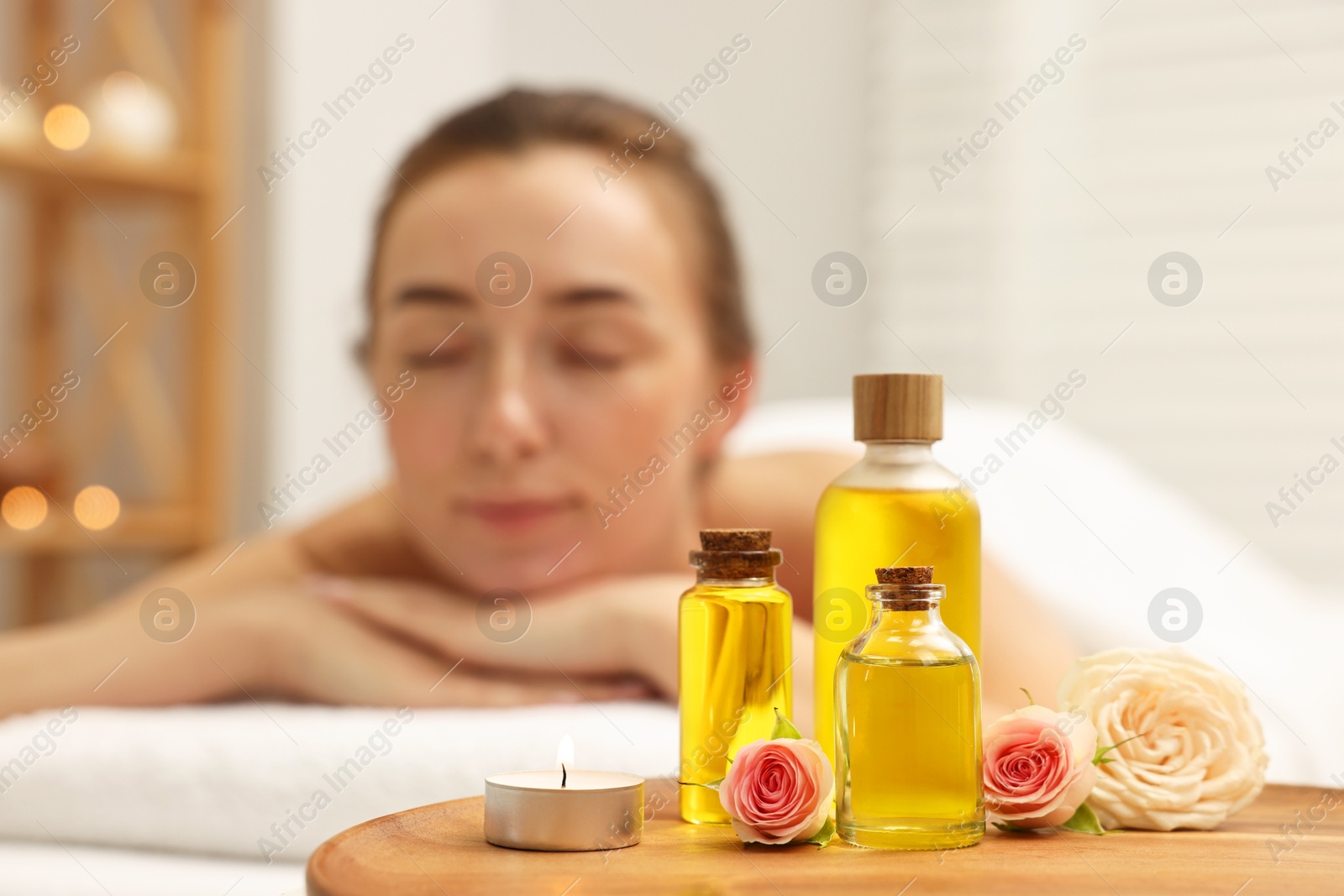 Photo of Aromatherapy. Woman relaxing on massage couch in spa salon, focus on bottles of essential oils, burning candle and rose flowers