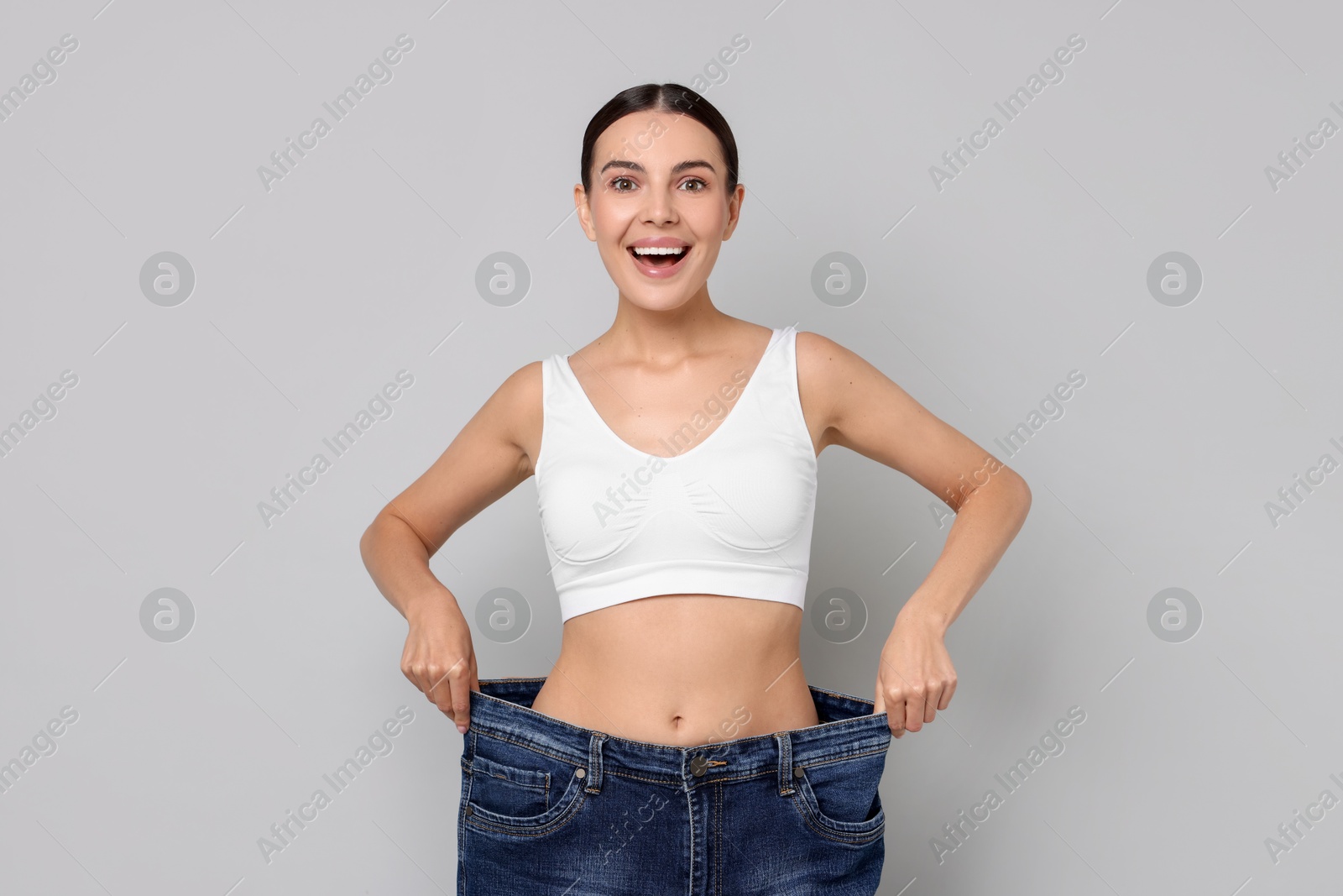 Photo of Diet and weight loss concept. Happy young woman in big jeans showing her slim body on light grey background