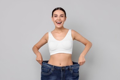 Diet and weight loss concept. Happy young woman in big jeans showing her slim body on light grey background