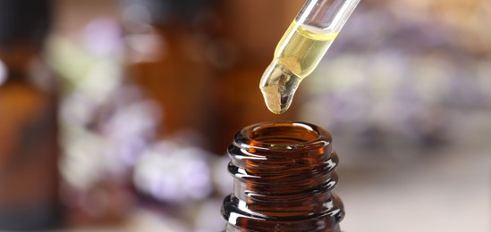 Photo of Essential oil dripping from pipette into bottle against blurred background, closeup