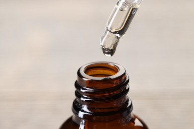 Photo of Essential oil dripping from pipette into bottle against blurred background, closeup