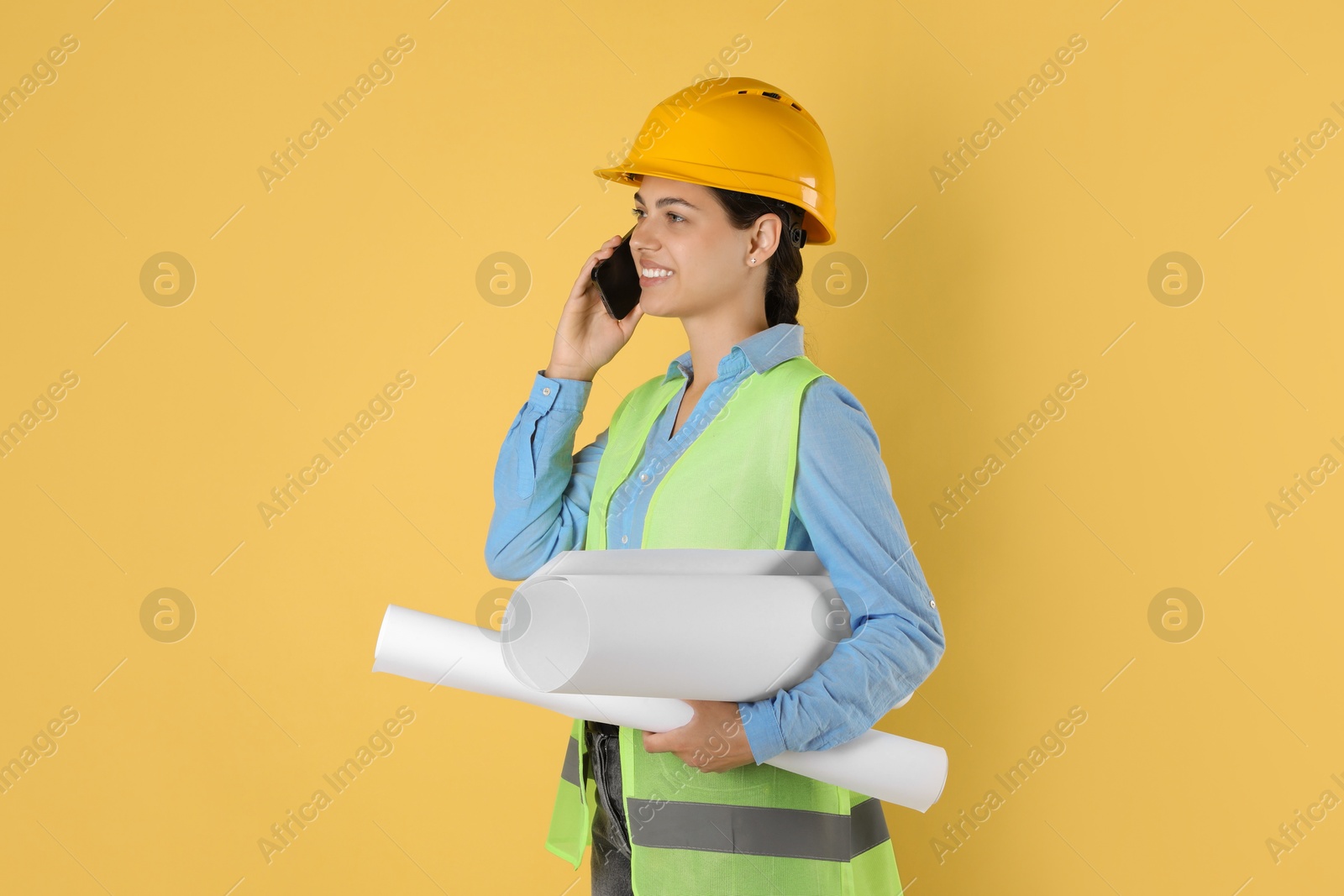 Photo of Engineer in hard hat with drafts talking on smartphone against yellow background