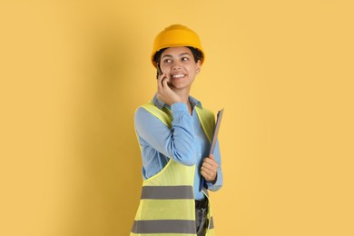 Photo of Engineer in hard hat with clipboard talking on smartphone against yellow background
